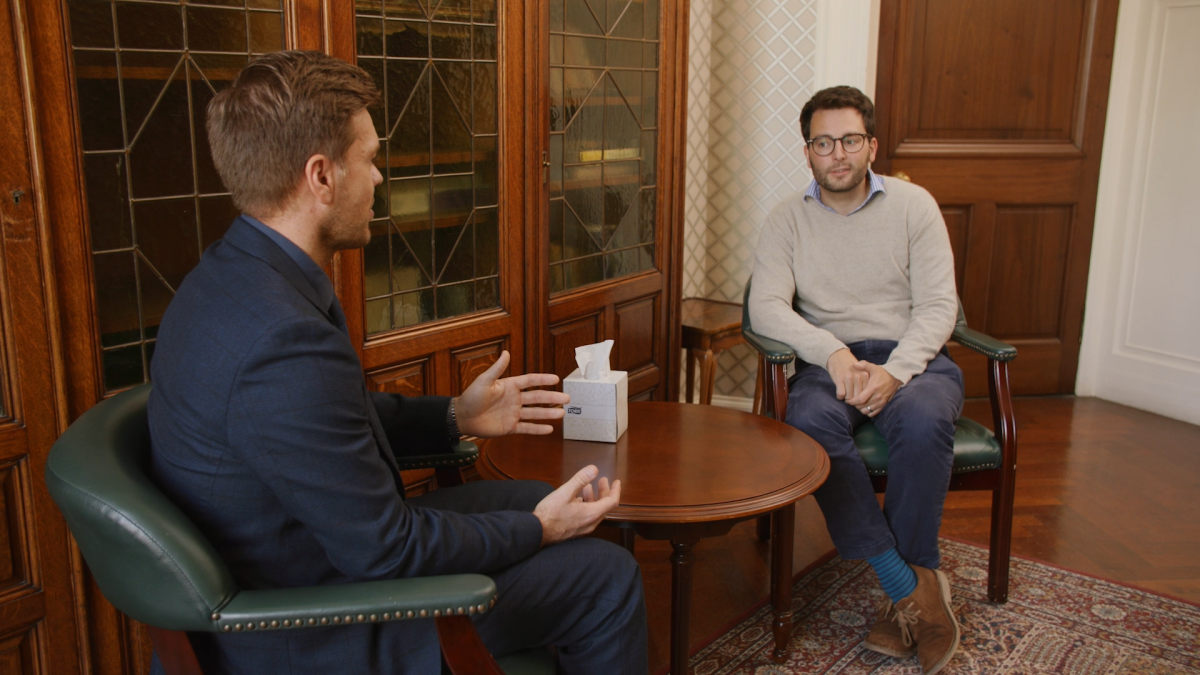 Two men sitting in a formal office setting having a professional conversation with a small round table between them holding a box of tissues elegant wood paneling and classic decor in the background